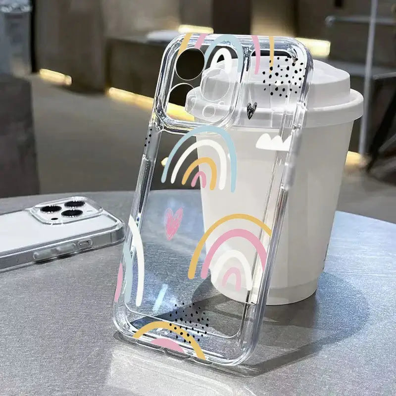 Transparent iPhone case with colorful heart and rainbow graphic on a table with a coffee cup in the background.